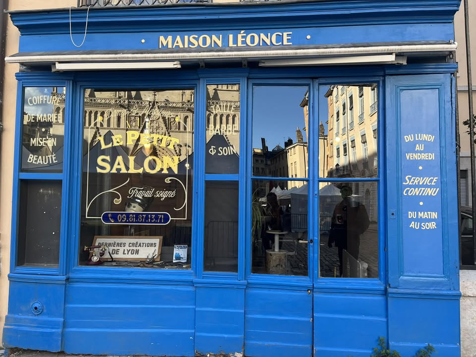 La devanture du Petit Salon, place Saint-Jean : façade bleu cobalt, enseigne dorée Maison Léonce, et sur la vitrine « Le Petit Salon, travail soigné », « coiffure de mariée, mise en beauté », « taille barbe et soin ».
