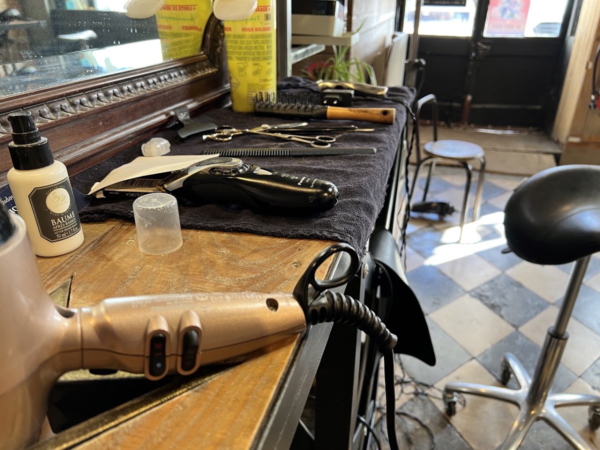 Le plan de travail en bois du salon : plusieurs paires de ciseaux, un peigne, une tondeuse, un rasoir coupe-chou à manche de bois et un baume après-rasage, avec le sol en damier noir et blanc derrière.